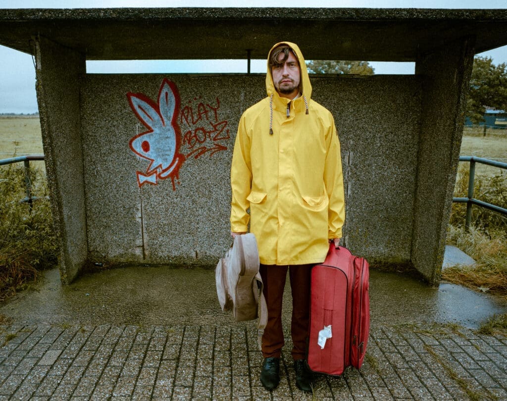 David J Kirchner steht in einem gelben Regenmantel an einer Bushatestelle. In der rechten Hand einen Gitarrenkoffern, in der linken einen Rollkoffer. Die Stimmung ist nass und regnerisch.