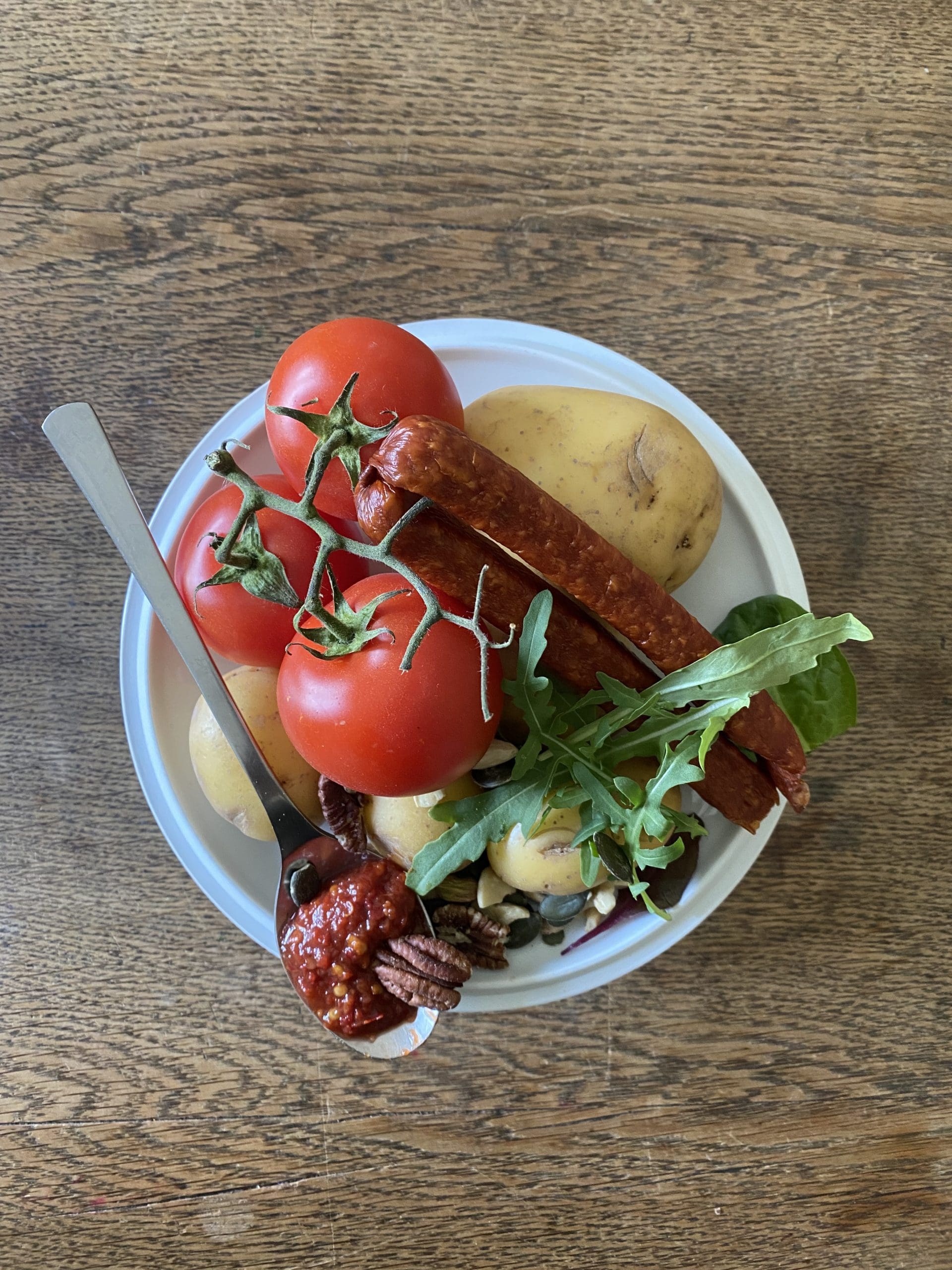 Auf dem Bild ist eine Schüssel mit Essen abgehandelt. In der Schüssel befinden sich Tomaten, rohe Kartoffeln, zwei Würste, etwas Salat, und wenige Nüsse. Auf der Schüssel liegt ein Löffel mit einer roten Paste und einer Nuss.